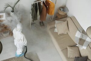 A person wearing a white protective suit and face mask using a fogging device to disinfect or fumigate a residential living space. The room features a beige couch, pillows, and a clothing rack, with visible mist spreading throughout, creating a clean and sanitized environment.